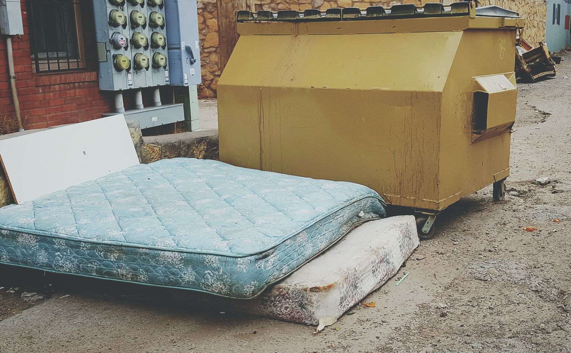 Dumpster and discarded mattress on a city sidewalk.