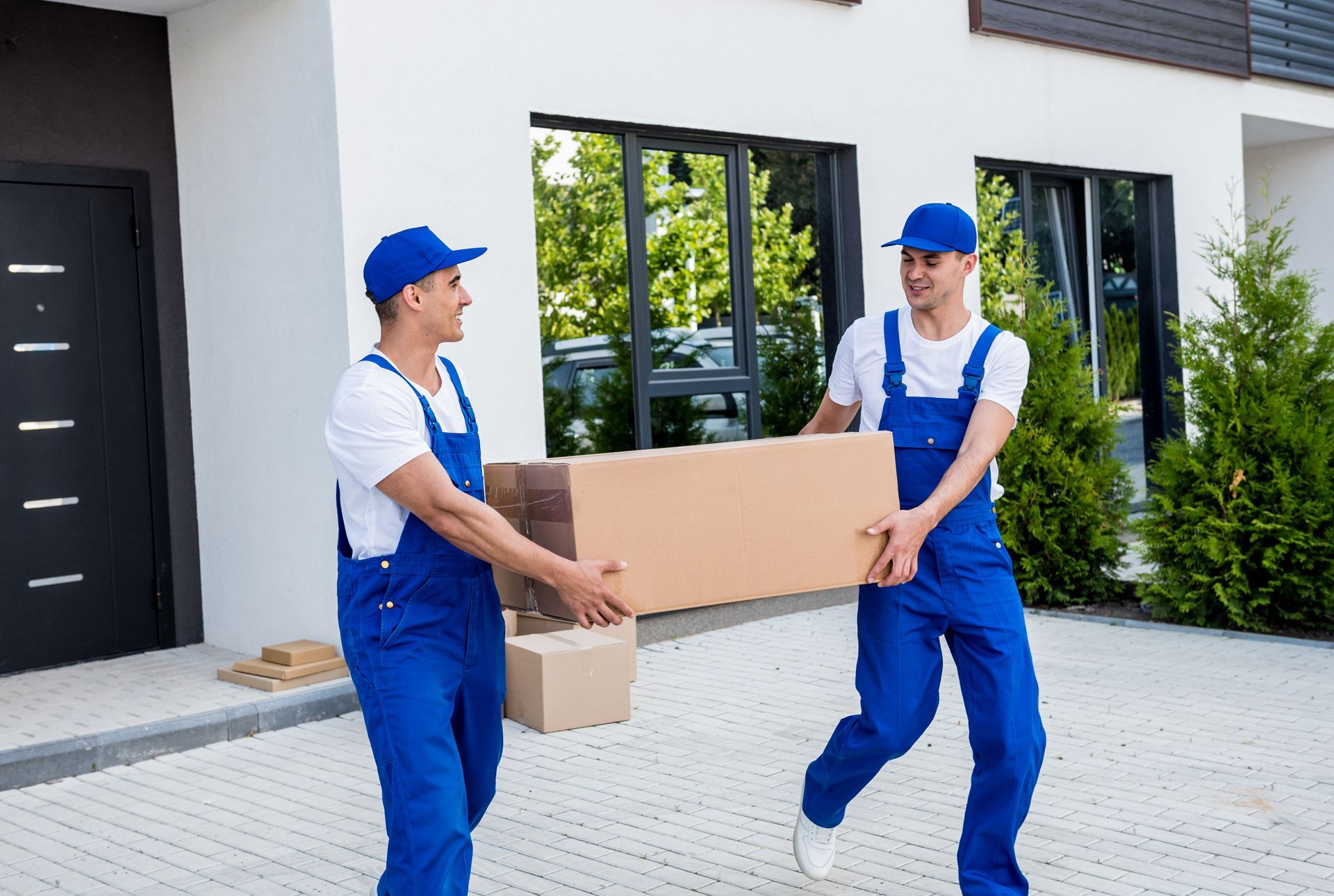 Two movers are carrying a cardboard box in front of a house.