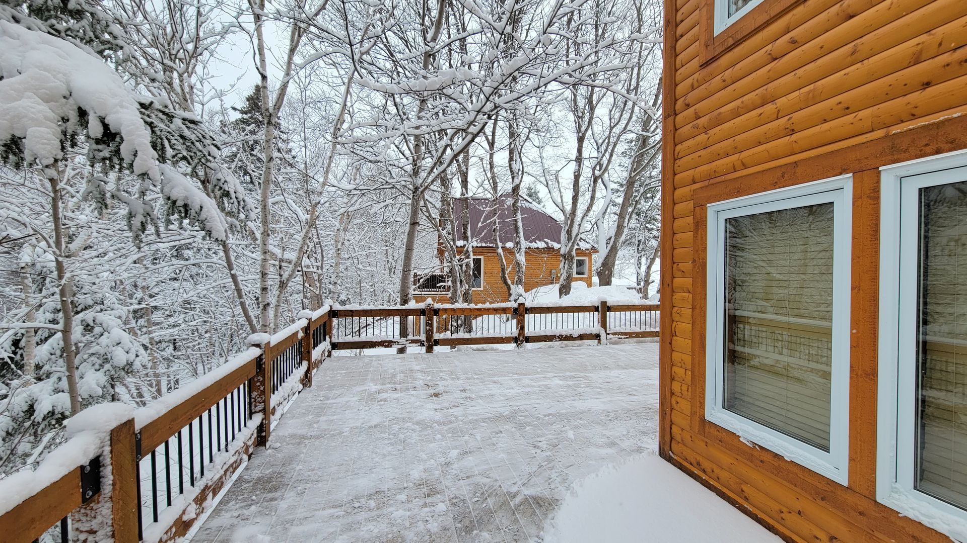 Snow-covered deck of a wooden house, overlooking a snow-covered forest and another house.
