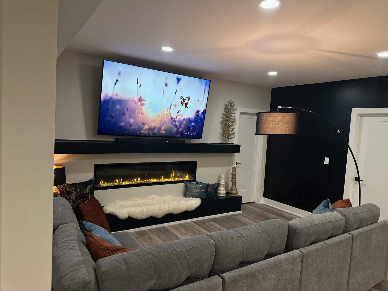 Modern living room with a gray sectional, fireplace, large TV, and black accent wall.