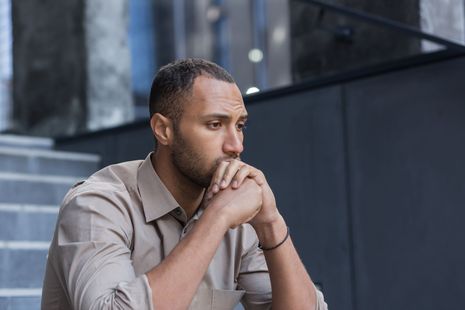 Man with dark hair and skin, resting his chin on clasped hands, looking down with a concerned expression