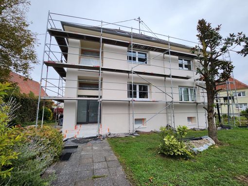 Des échafaudages entourent une maison de trois étages en cours de rénovation, avec un enduit blanc fraîchement appliqué et des fenêtres visibles.