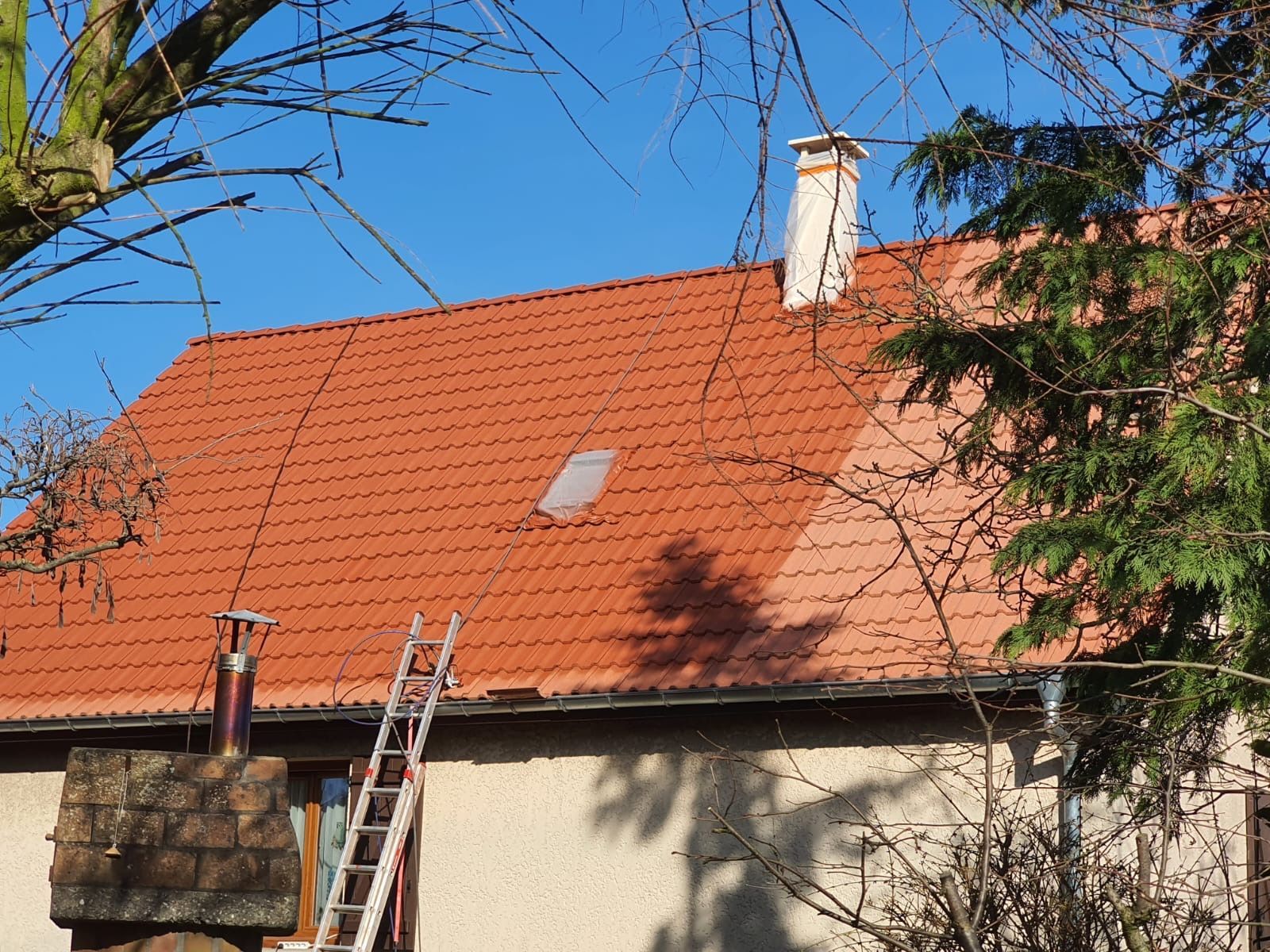 Toit de tuiles rouges avec cheminée, échelle et clairière. Le tout se détache sur un ciel bleu bordé d'arbres.