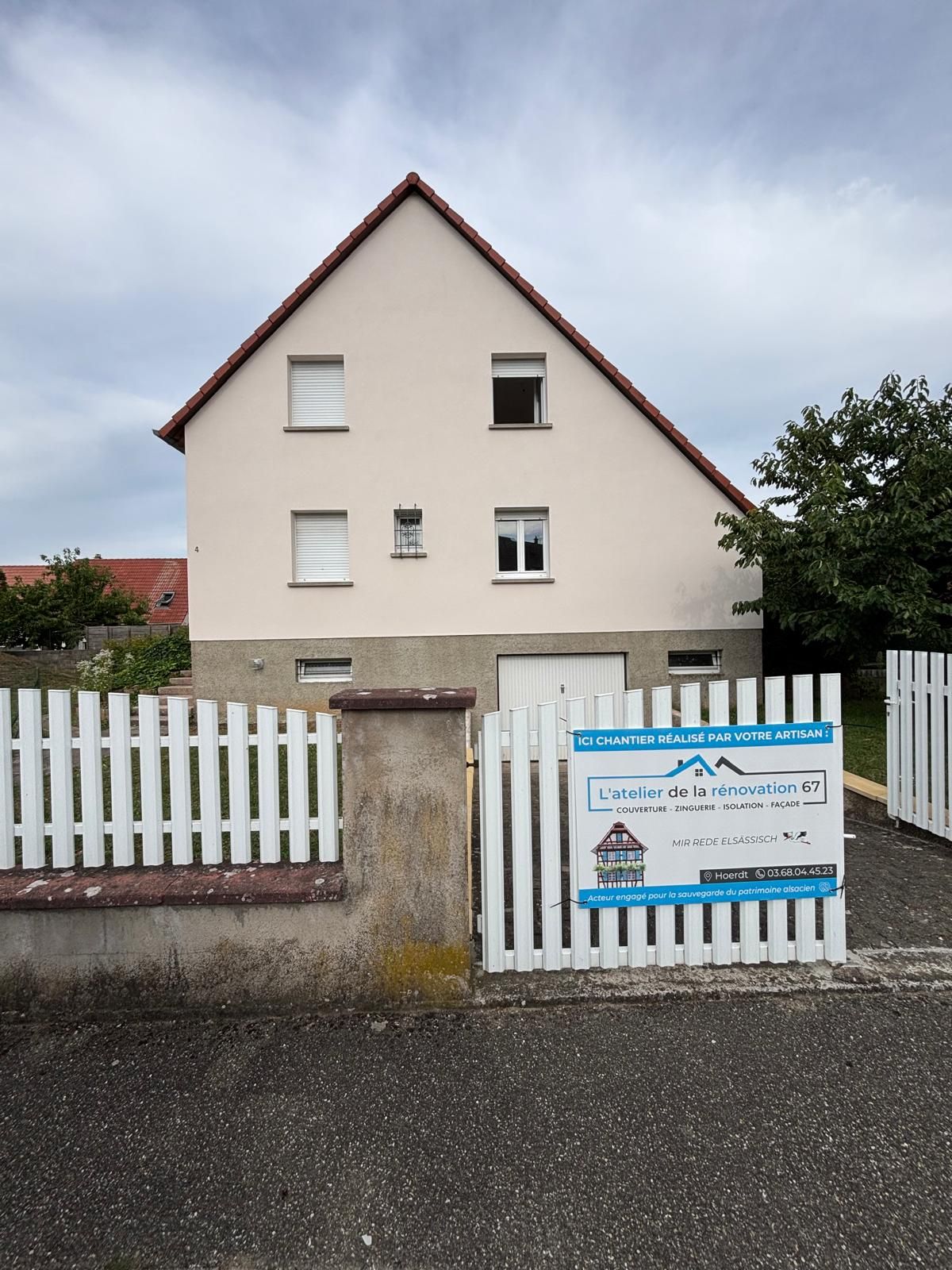 Maison beige à deux étages avec un toit rouge, une clôture blanche et un panneau. Ciel nuageux en arrière-plan.