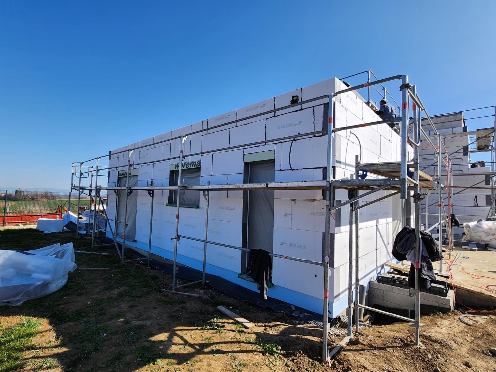 Bâtiment en construction, recouvert de panneaux isolants blancs et entouré d'échafaudages. Ciel bleu.