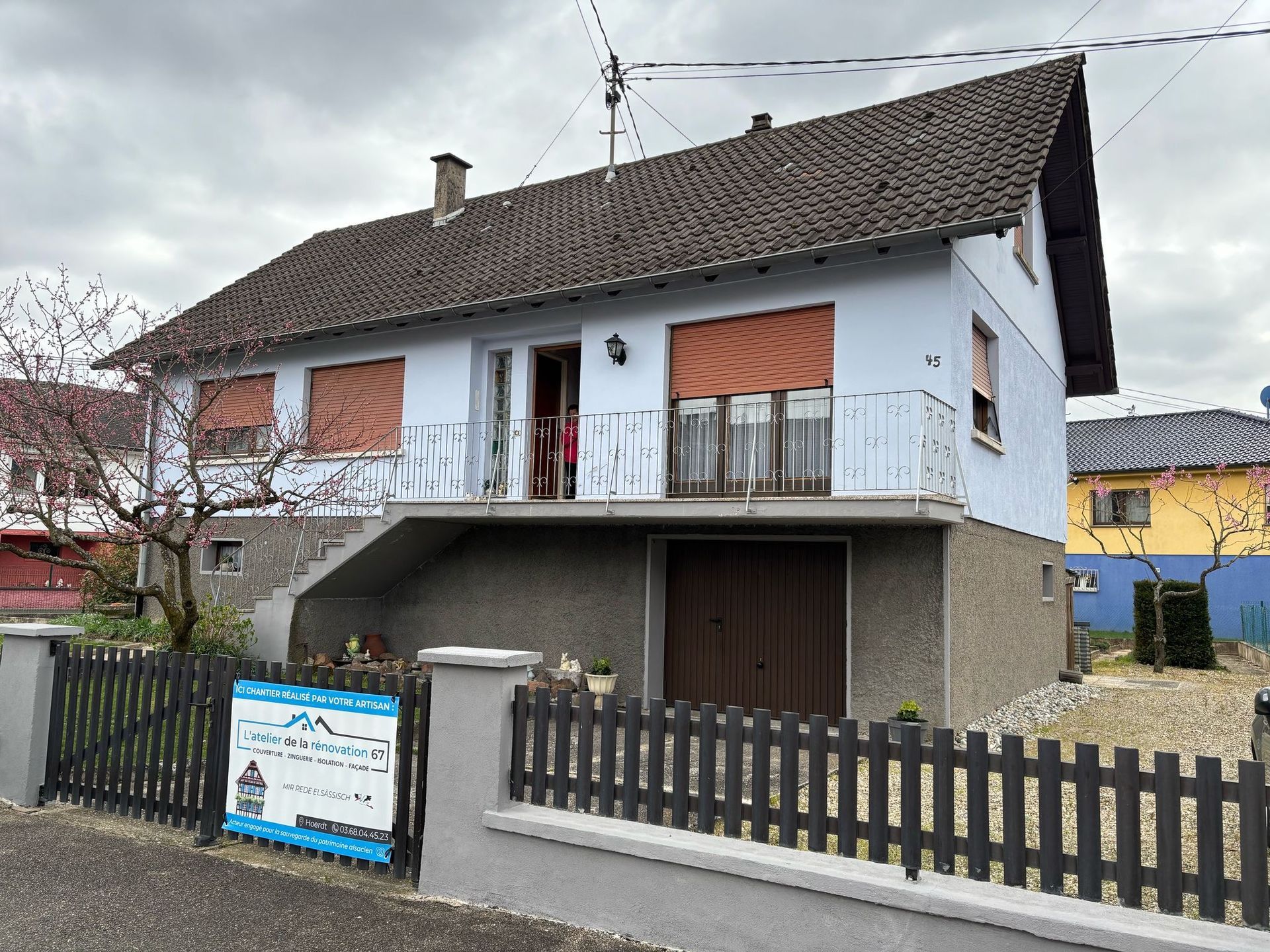 Maison bleue à deux étages avec toit marron et garage. Balcon avec stores rouges. Clôture noire à l'avant.