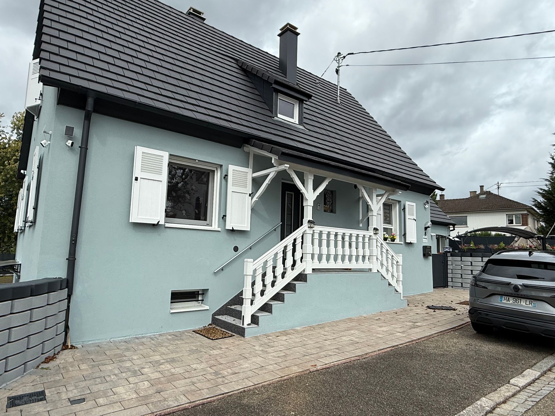 Maison bleu clair avec boiseries blanches, porche et volets, toit gris. Escalier et allée en pierre. Voiture garée à droite.