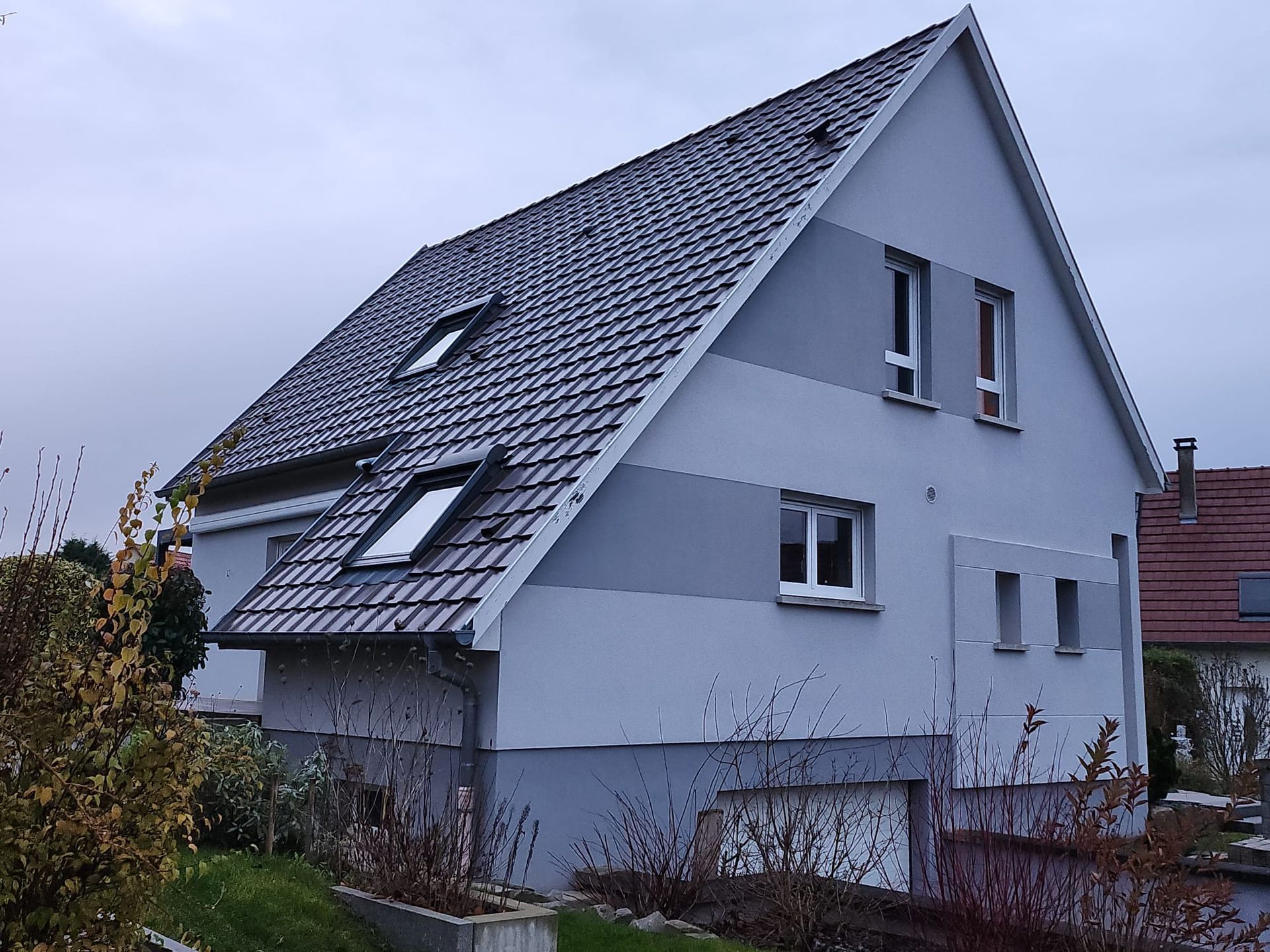 Maison à deux étages avec façade grise et blanche, toit sombre et petites fenêtres. Ciel couvert.