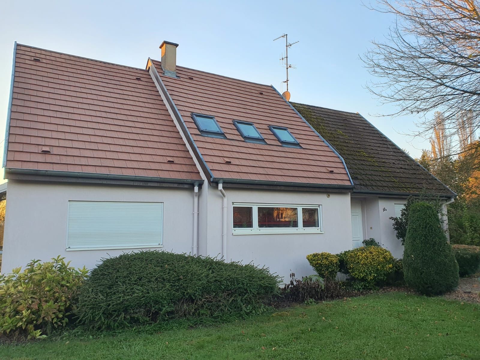 Maison rose au toit de tuiles rouges, petites fenêtres et verdure environnante sous un ciel bleu.