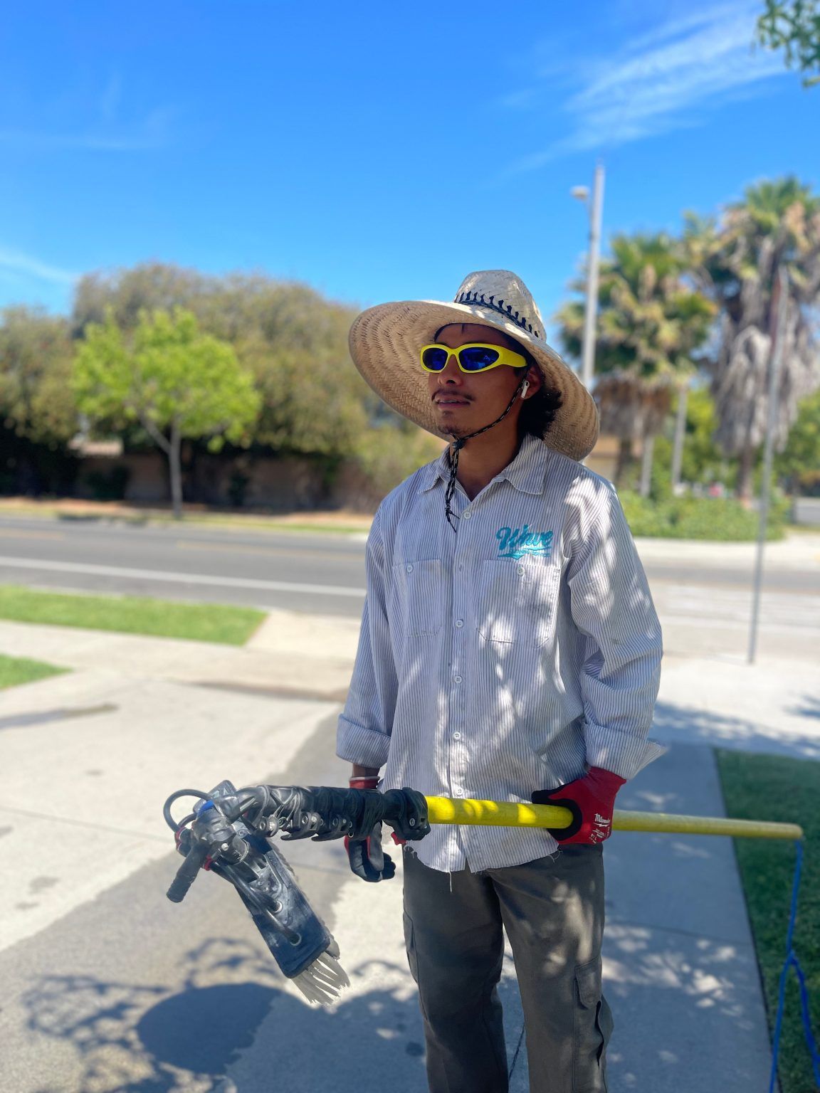 Person in straw hat and sunglasses holding a tool, standing outdoors near a sidewalk on a sunny day.