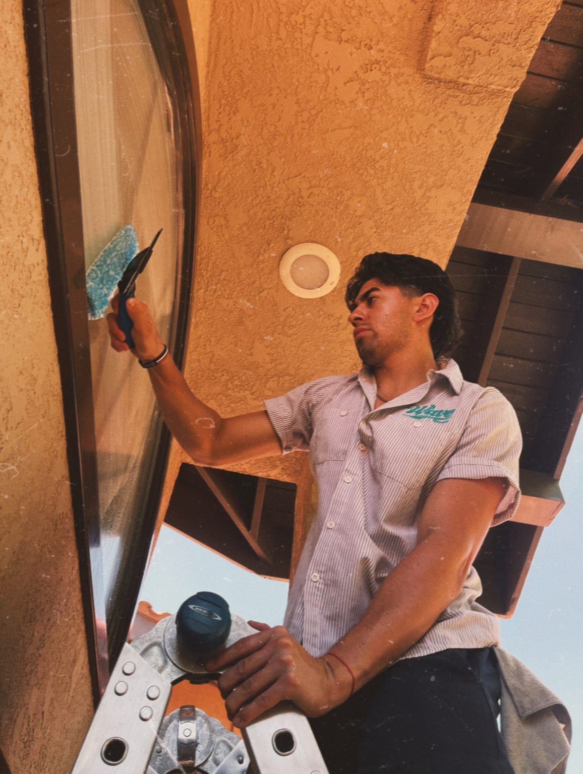 Man cleaning window with a squeegee while standing on a ladder. Building exterior visible.
