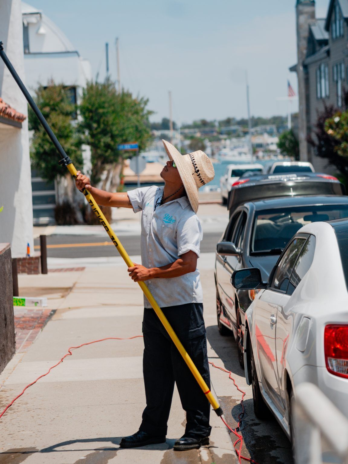Man in straw hat power washing a building's exterior on a sunny day near parked cars and water.