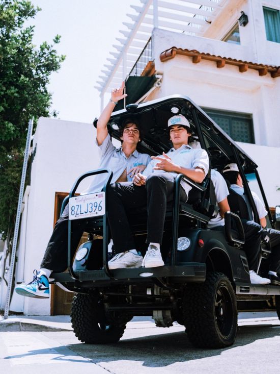 People in a modified golf cart, one waving, near a white building with a ladder.