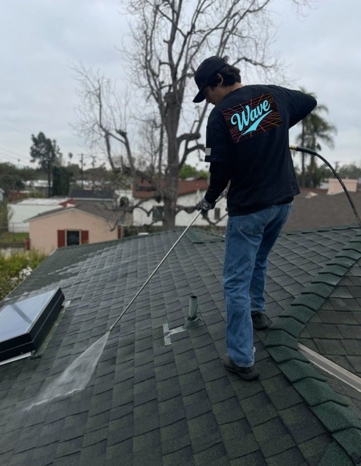 Person pressure washing a roof gutter. Water spraying, sunlit exterior, blue sky, trees.