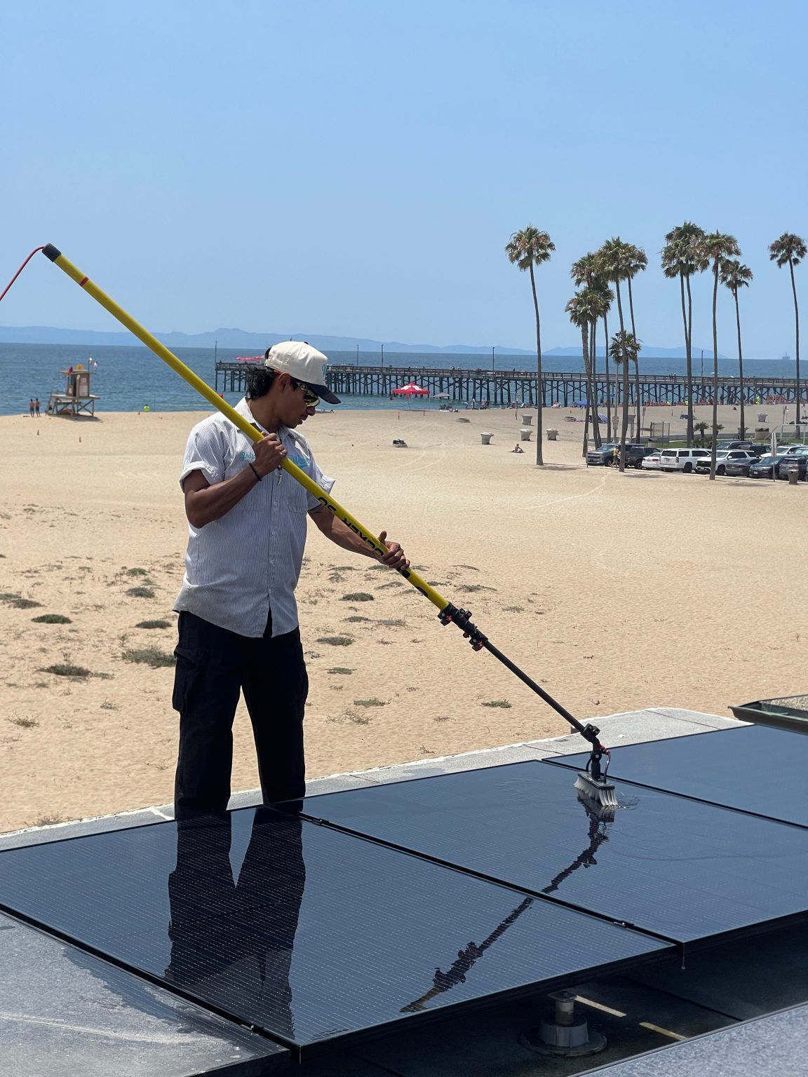 Man cleaning solar panels on a rooftop with beach and pier in the background. Bright sunny day.