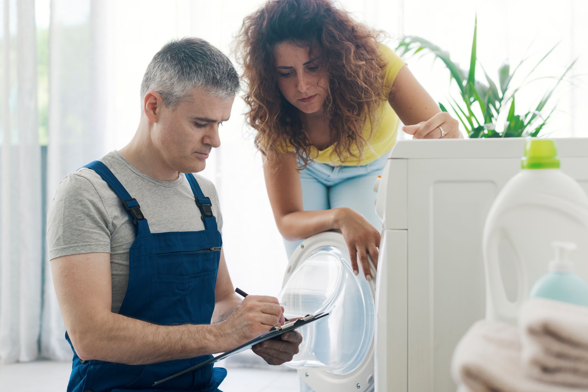 Man in overalls writes on clipboard, woman watches near a washing machine.
