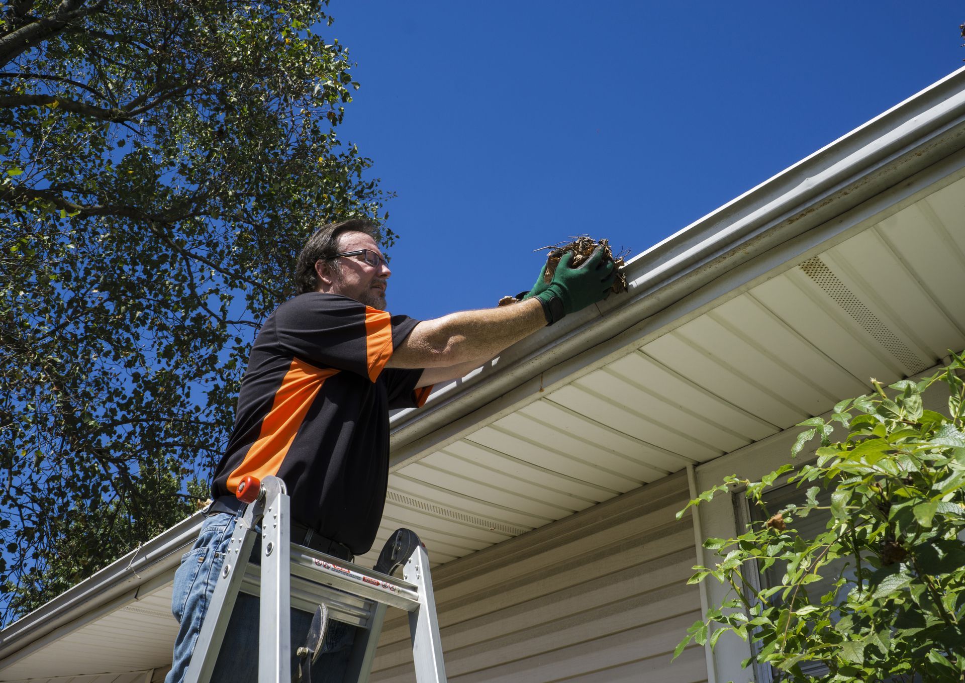Man on ladder cleaning a gutter with gloved hand against blue sky.