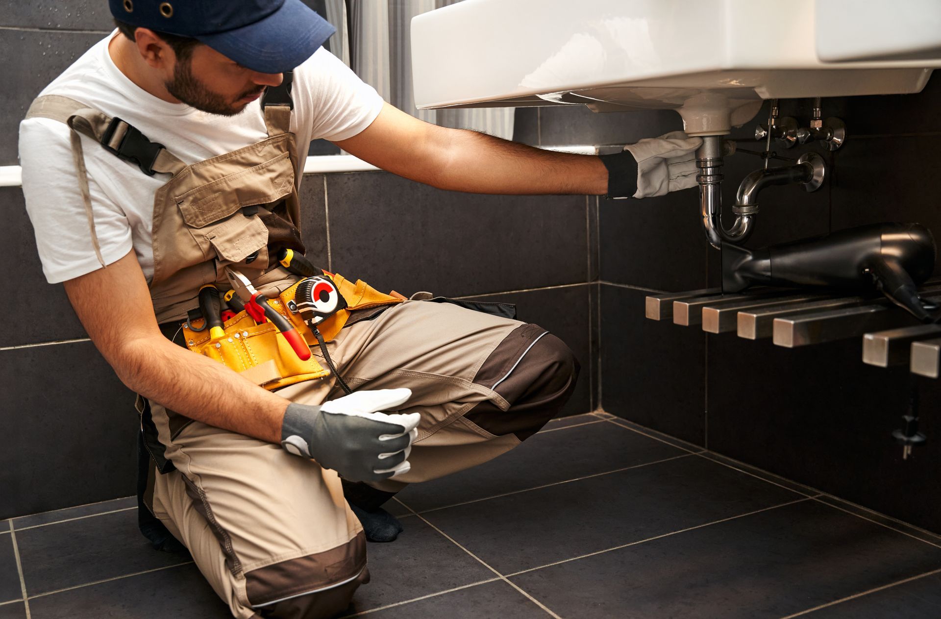 Plumber kneeling by a bathroom sink, working on the pipes. Dark tiles and a tool belt are visible.