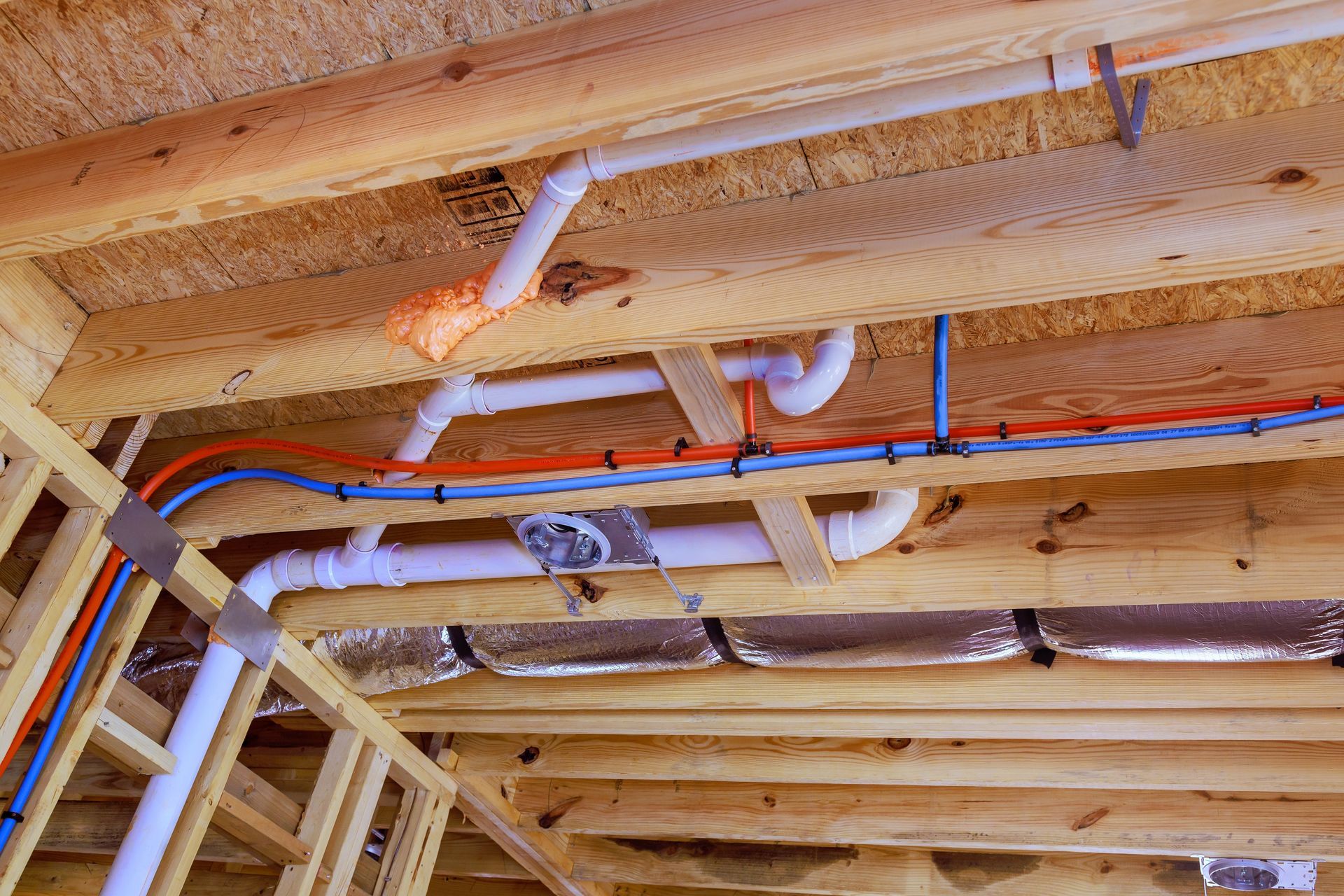 Exposed ceiling framing with white PVC pipes and red/blue plumbing lines in a house under construction