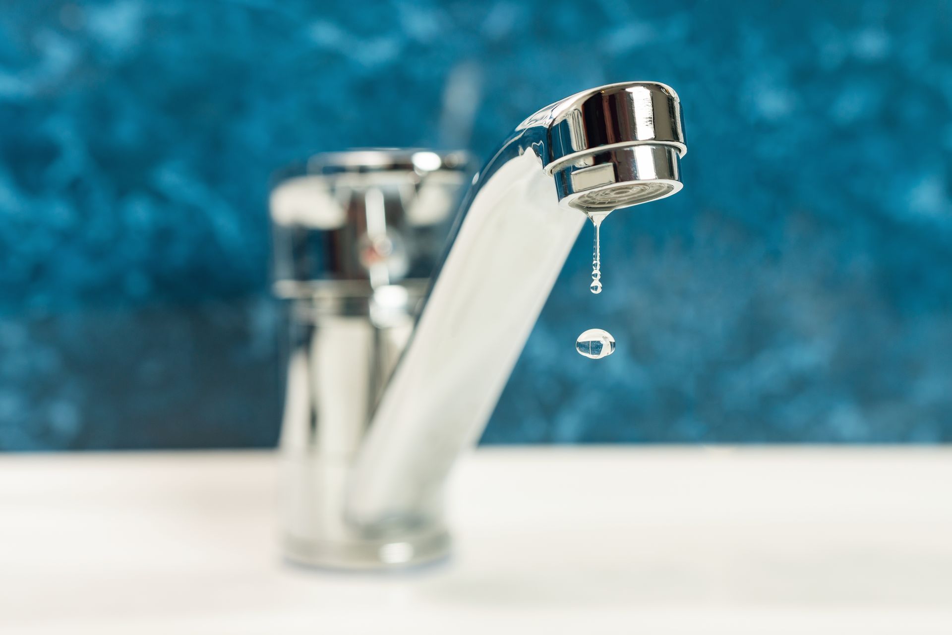 A chrome faucet with water dripping, close-up view. Blue background, set on a white sink.