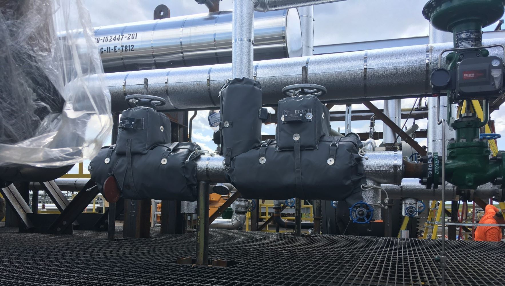 Industrial piping and black valve assemblies on a grated platform, with workers in safety gear nearby