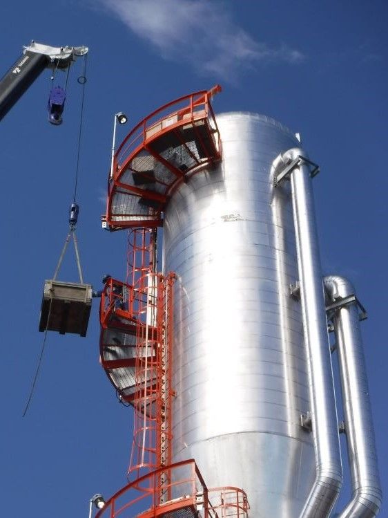 Industrial crane lifting a metal container beside a tall silver tank with red stairs and blue sky