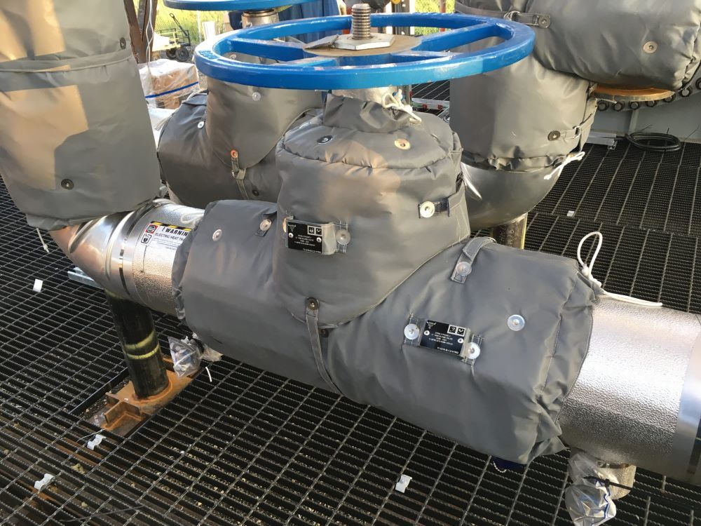 Insulated industrial pipes and valves with gray covers and a blue handwheel on a metal grating floor