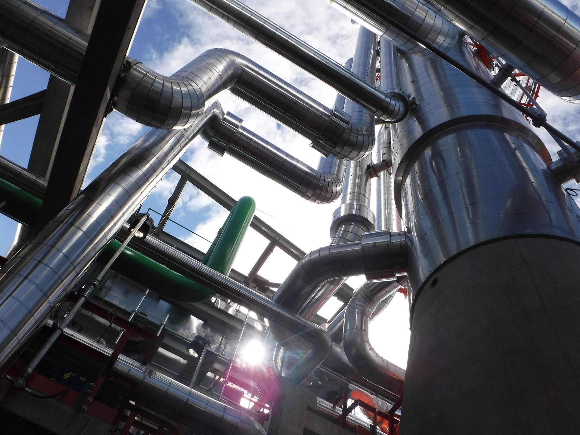 Shiny metal industrial pipes and tanks crisscrossing against a bright sky in a factory setting