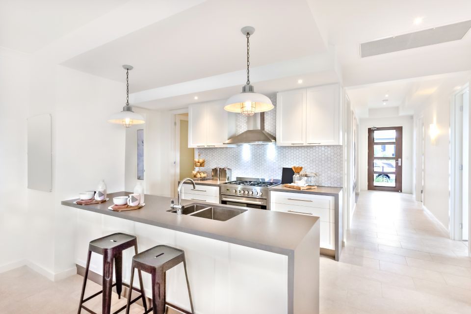 Cucina moderna e luminosa, di colore bianco, con isola centrale, lampade a sospensione, sgabelli da bar ed elettrodomestici in acciaio inossidabile.
