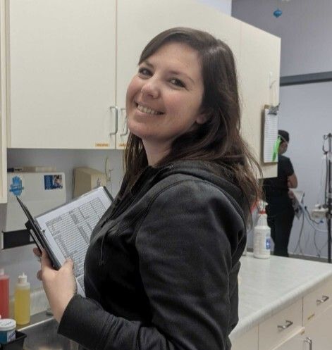 Woman in black jacket smiles, holds clipboard, standing in an office with a person blurred in the background.