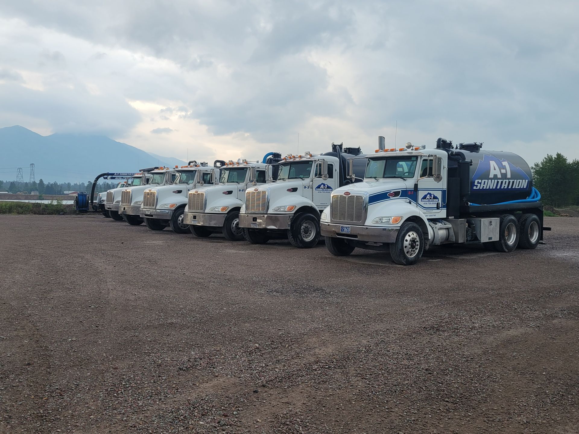 A line of white tanker trucks with blue and white logos parked on gravel. Mountains in the background.