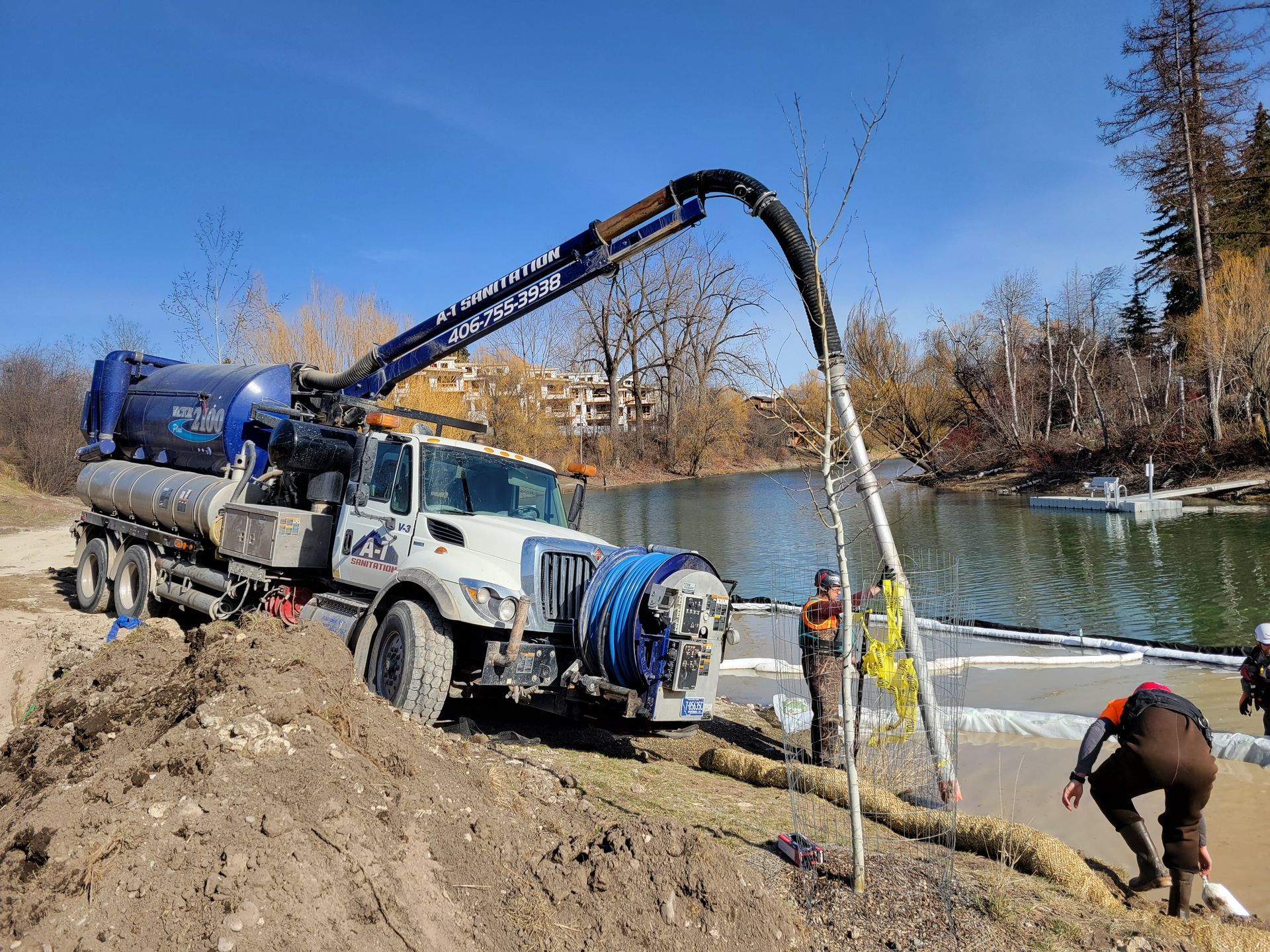 A large truck removing debris from a pond with workers in waders, on a sunny day.