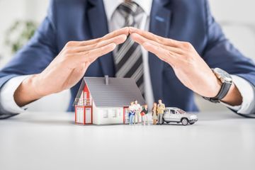 A man in a suit and tie is covering a house and a car with his hands.