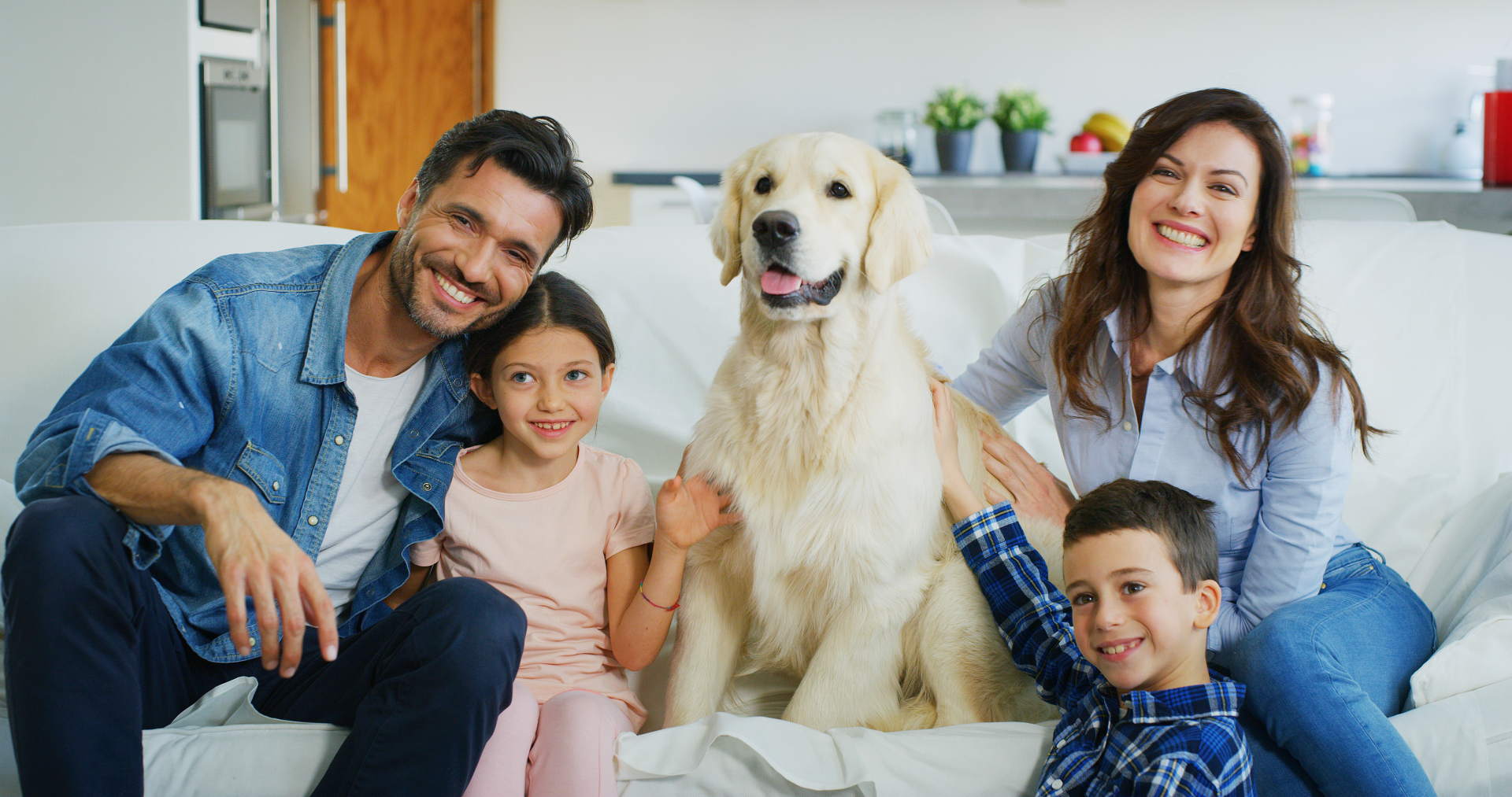 Family of five, including a golden retriever, smiles on a white couch in a home setting.