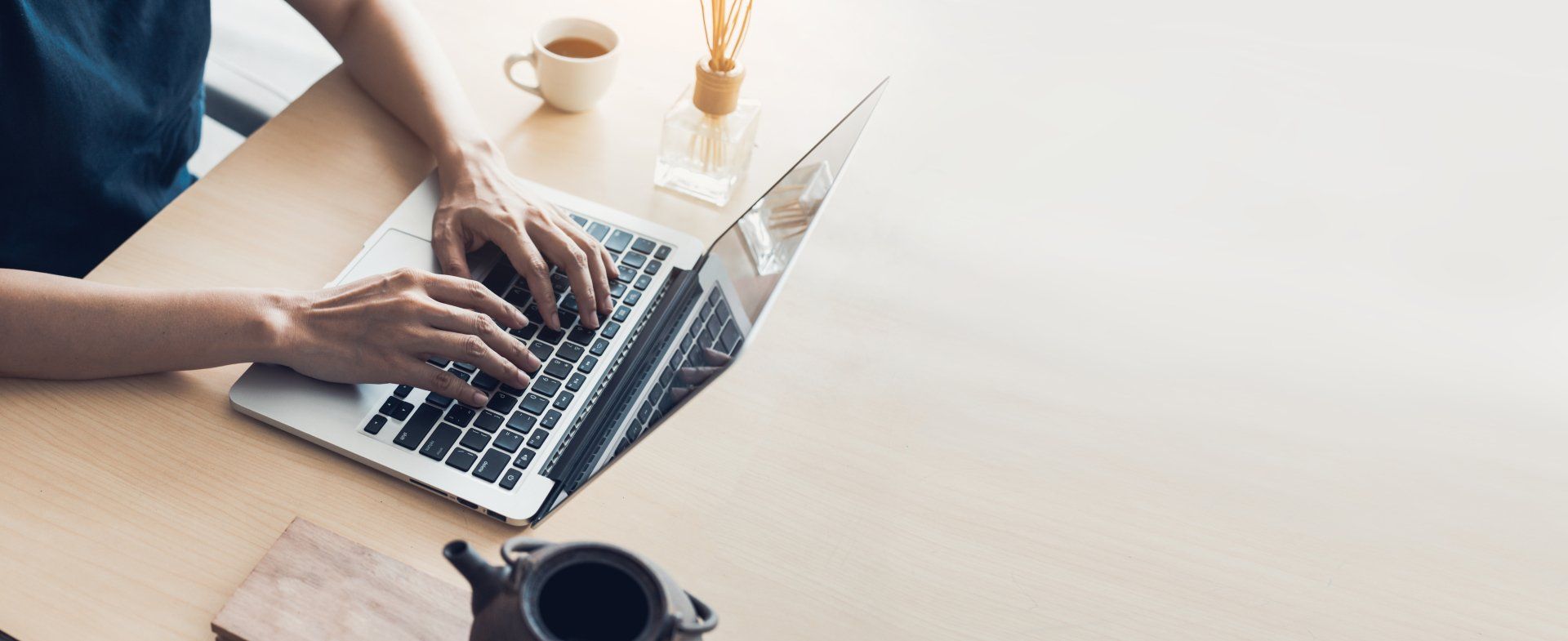 Person typing on a laptop on a wooden desk, with a cup of tea and a glass vase nearby.