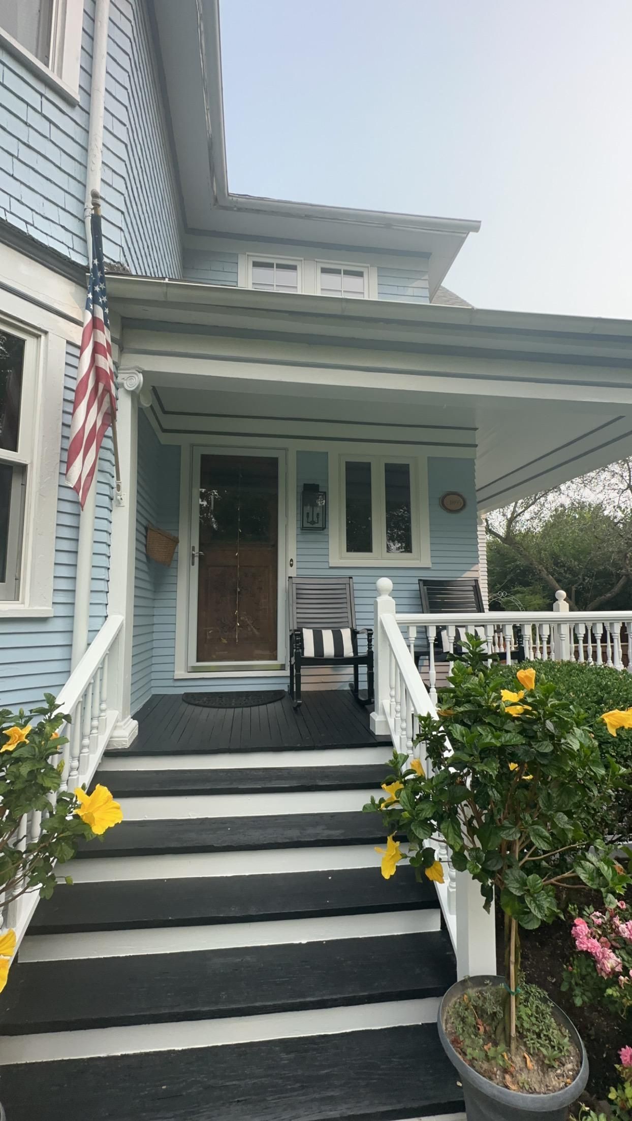 Blue Victorian house with front porch, American flag, and potted flowers.