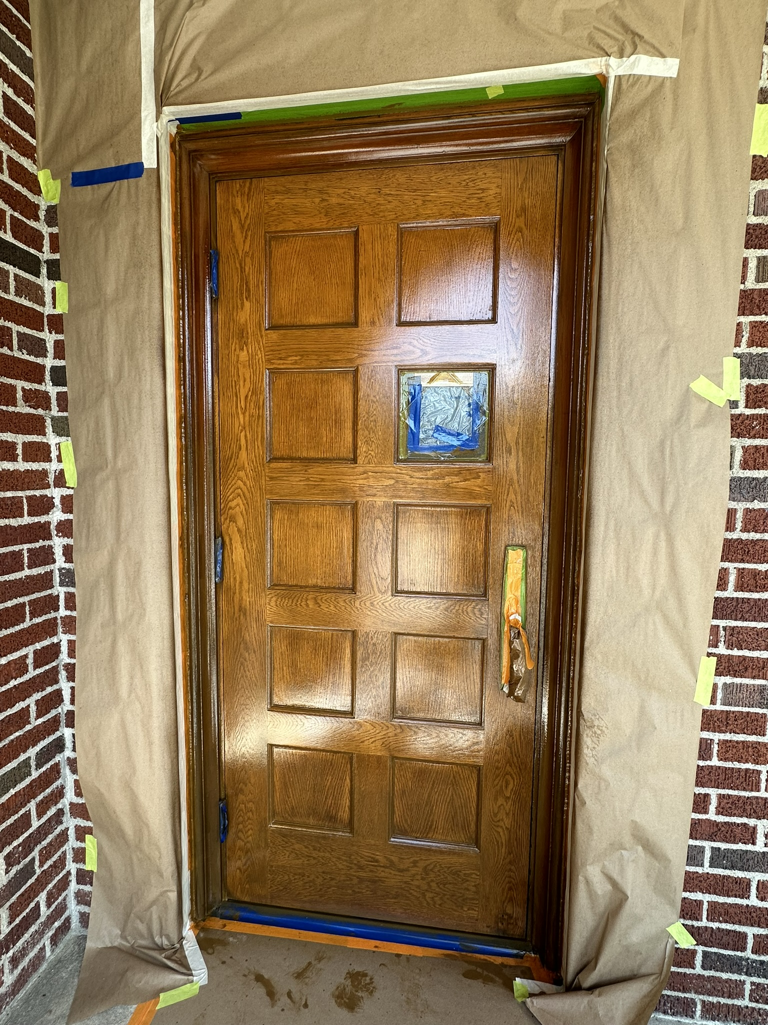 Wooden door, stained with brown finish, covered with protective paper and tape, against brick wall.