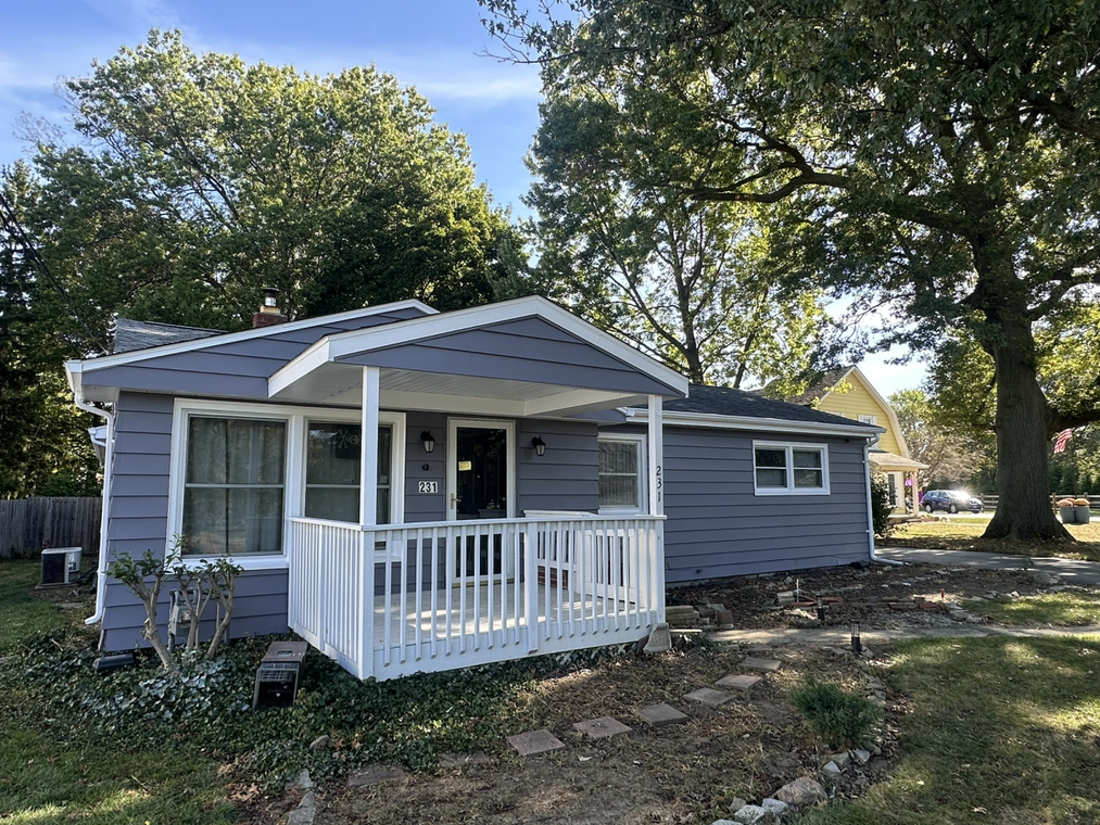Gray house with white porch, in a yard with trees under a blue sky.