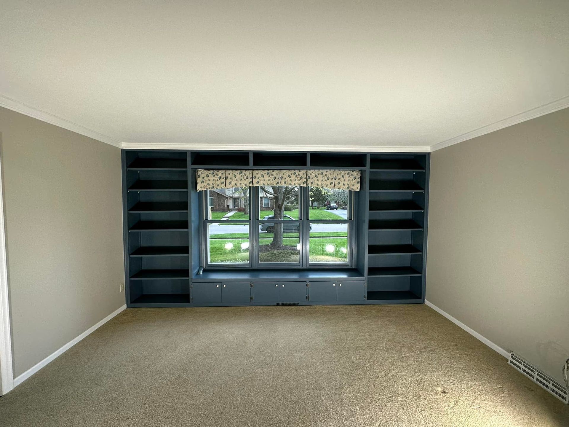 Empty room with built-in dark blue bookshelves around a window, tan walls, and beige carpet.