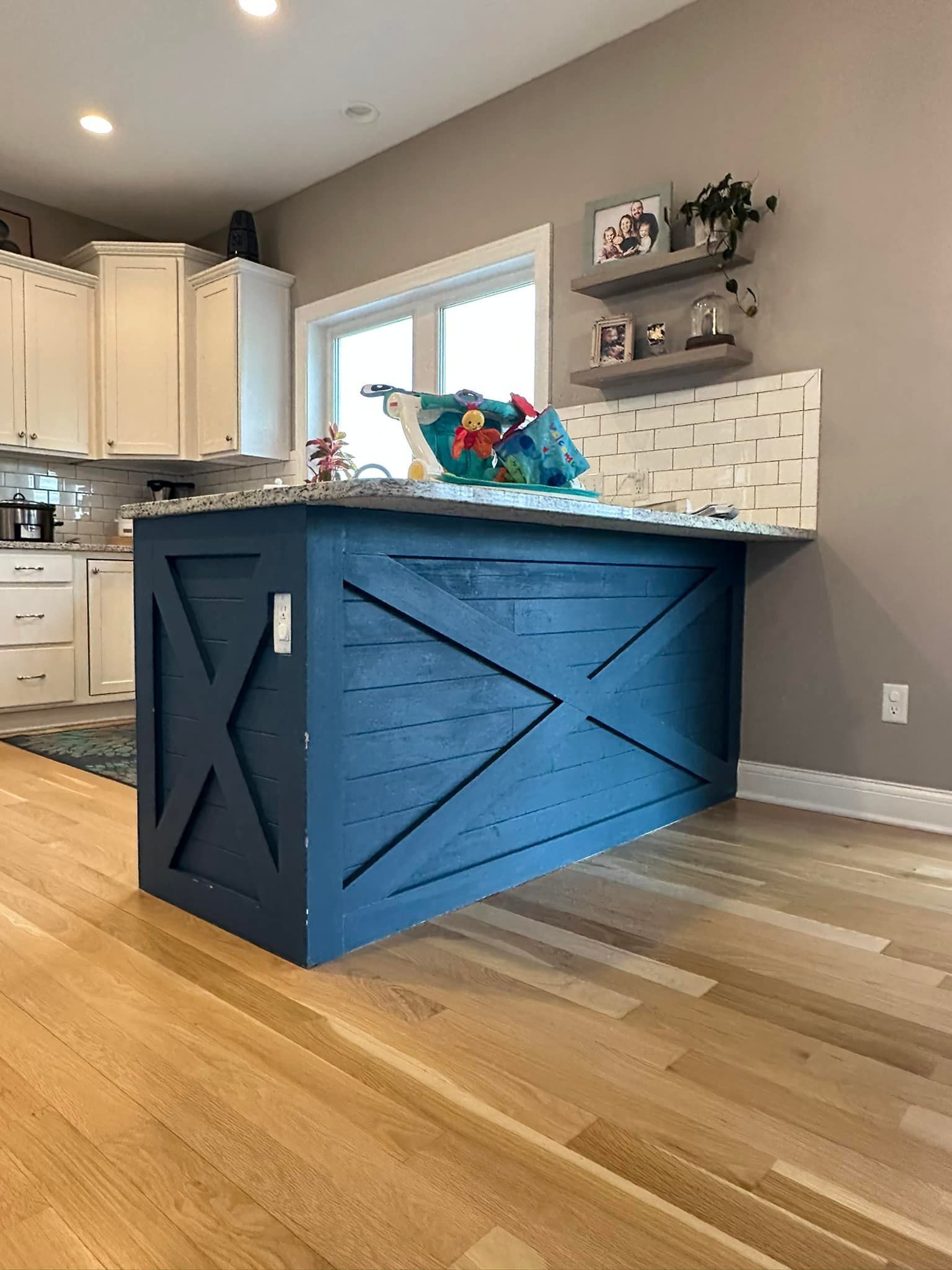 Blue farmhouse kitchen island with an X design, granite countertop, white cabinets, and wood flooring.