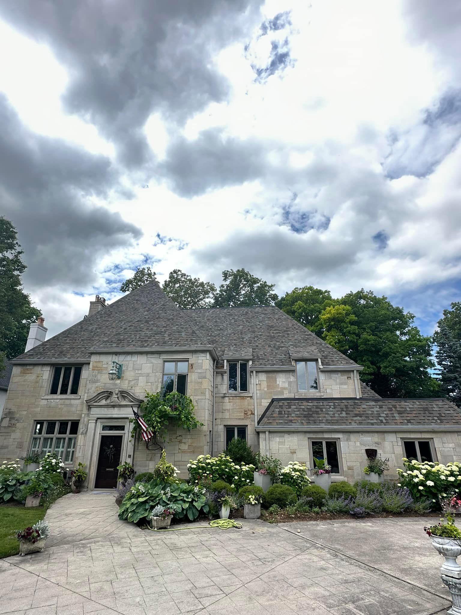 Stone house with gray roof, front porch, and flowers under cloudy sky.