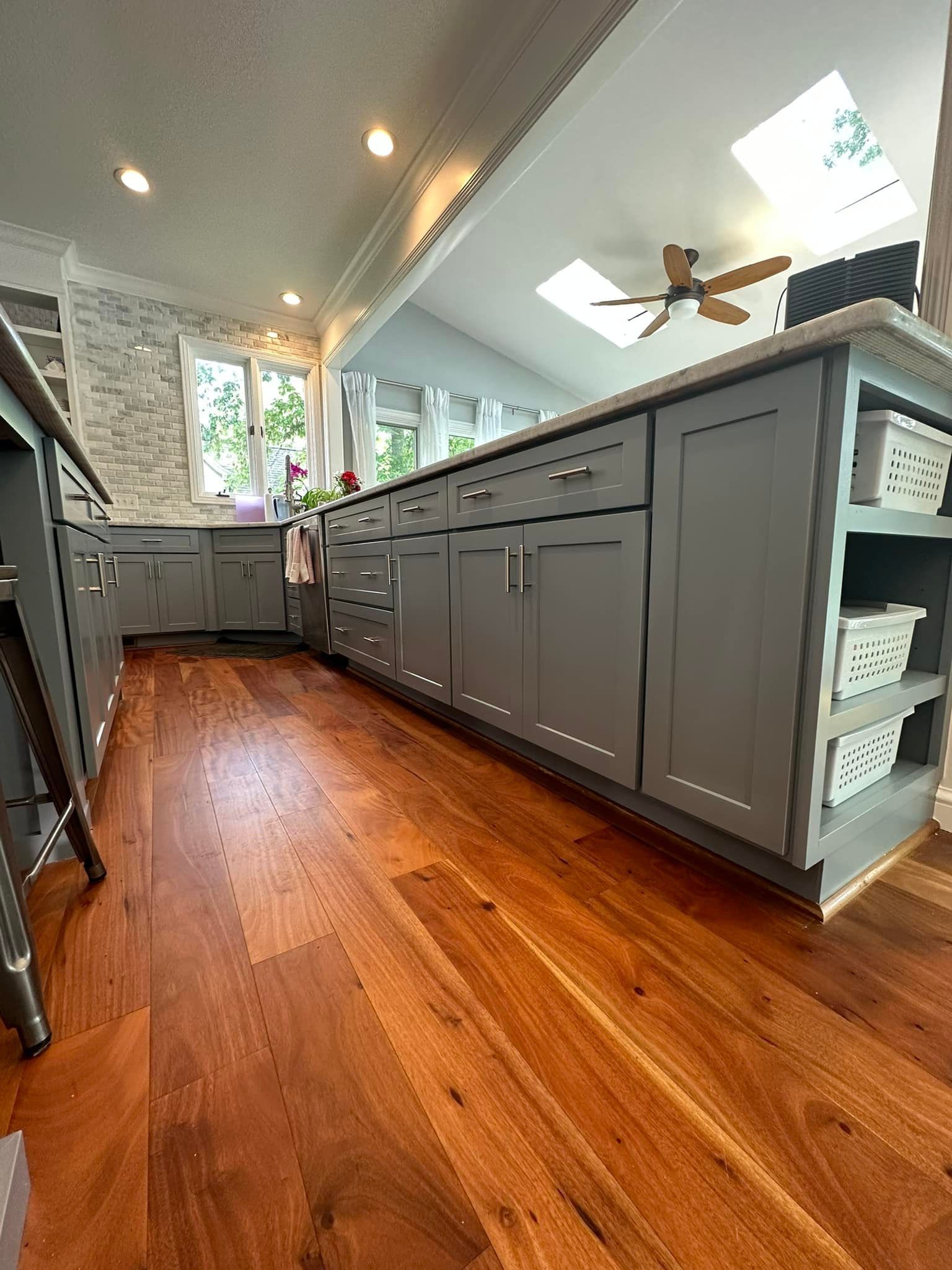Wooden floor, gray kitchen cabinets, island with open shelves, skylights, and a ceiling fan.