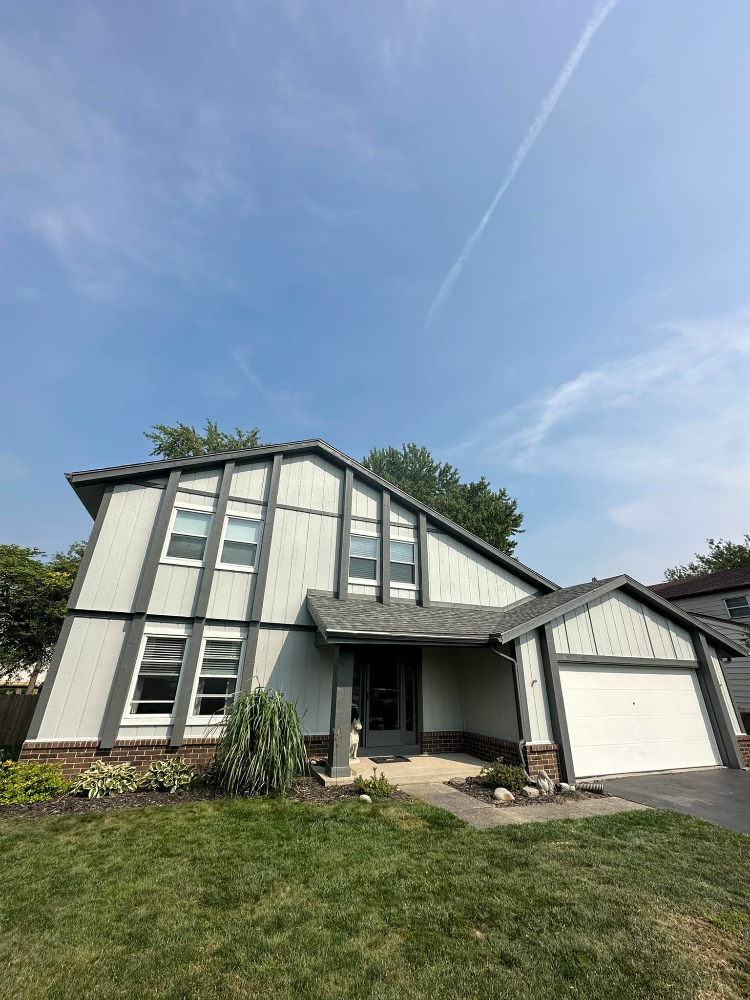 A two-story house with light gray siding and a gray roof sits under a blue sky. A garage is on the right.