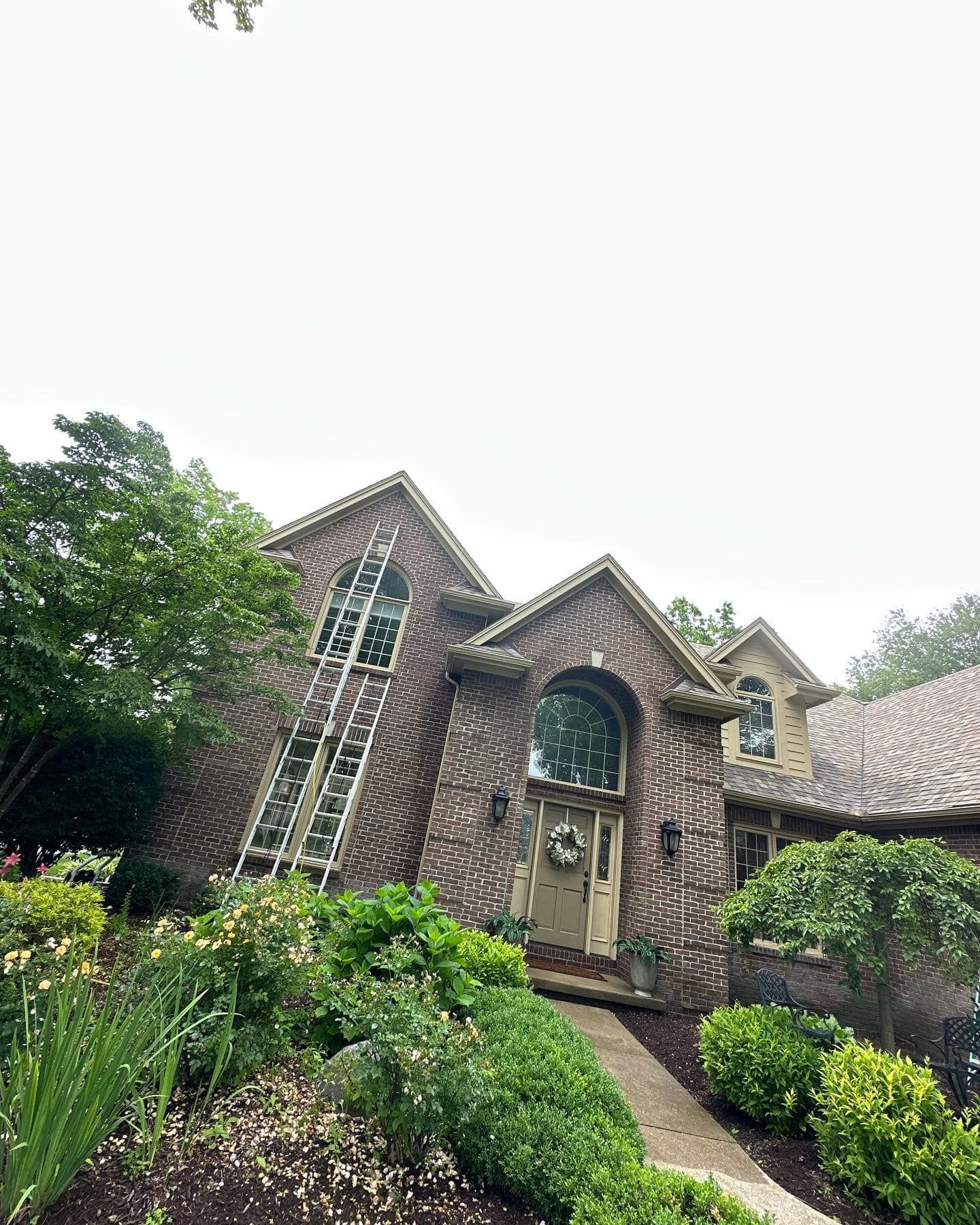 Brown brick house with arched window and doorway, surrounded by green bushes and trees.