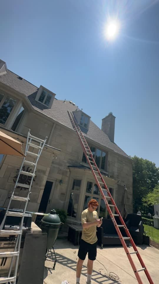 Person with phone near long ladder leaning against house with a gray roof, sunny day.