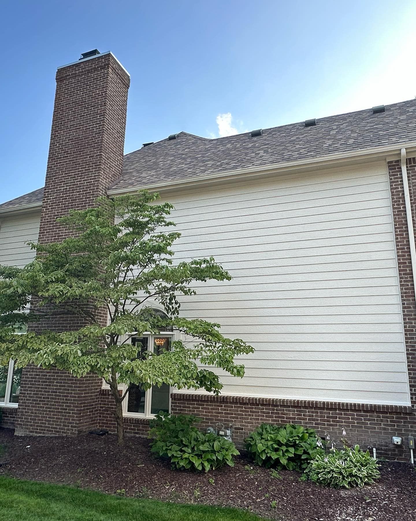 Side view of a house with brick chimney, white siding, and a small tree in front.
