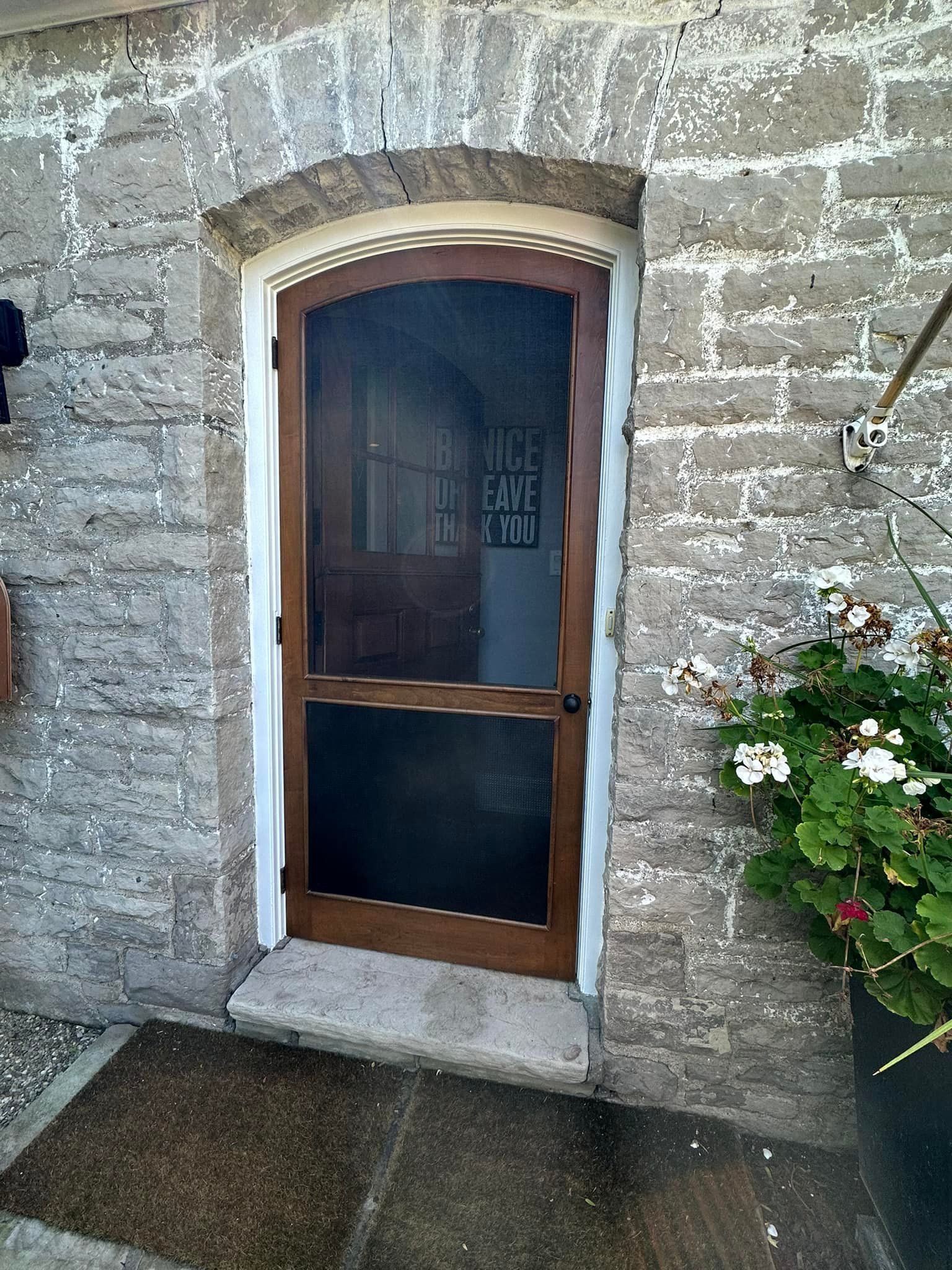 Wooden screen door with arched top in stone building. Geraniums in pot to the right.