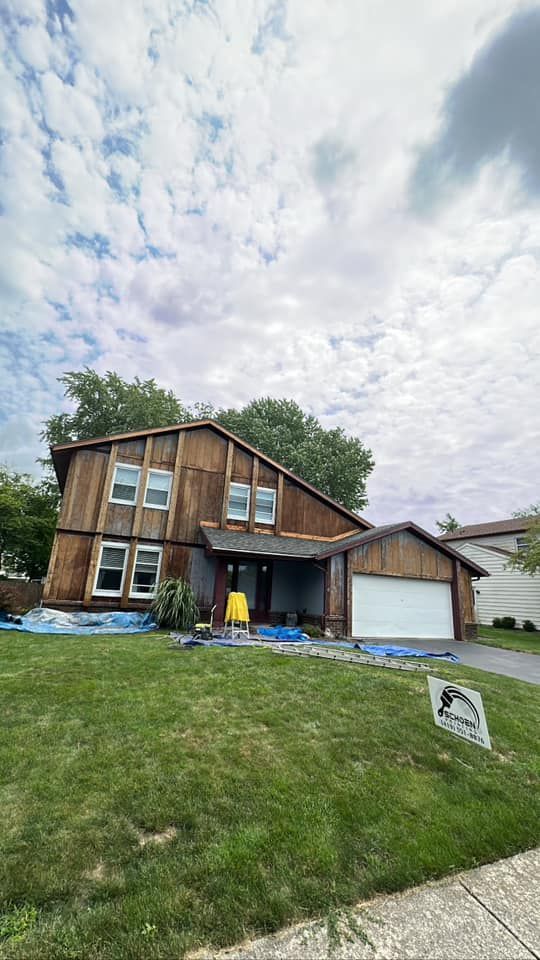 House under construction, wood siding removed, revealing wooden frame, blue tarp on lawn, cloudy sky.