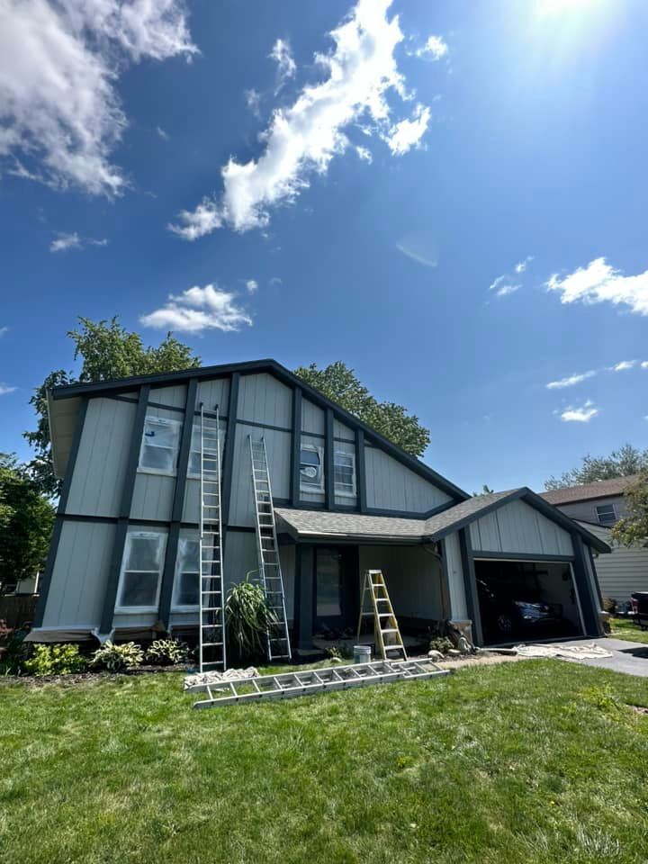 House exterior being painted, blue siding, white windows, ladders. Sunny day, blue sky, green grass.