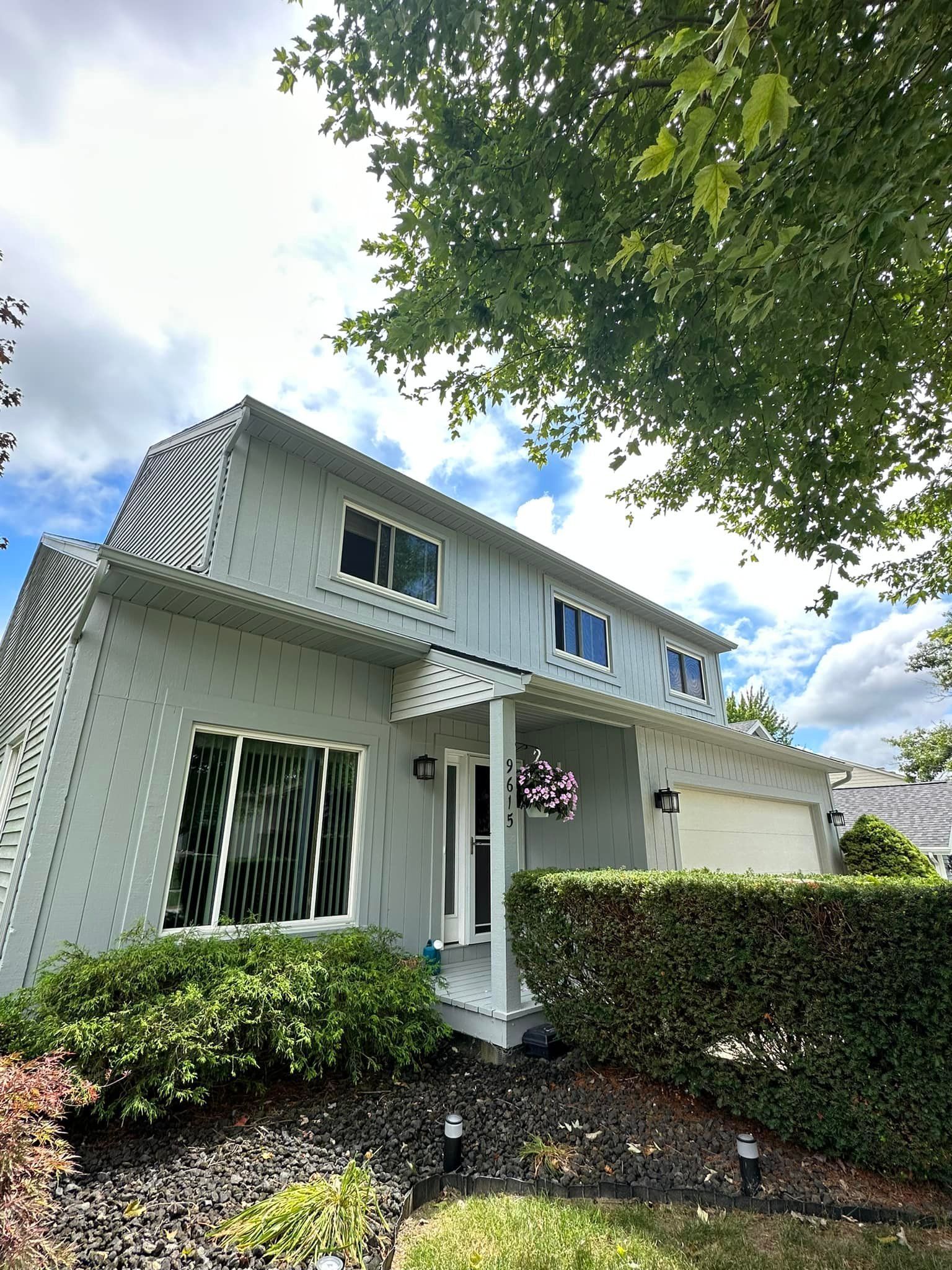 Two-story house with gray siding, white trim, and a green hedge in front of a blue sky.